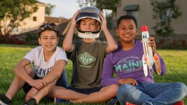 Three kids sitting on grass, playing with a toy rocket, enjoying a sunny day.